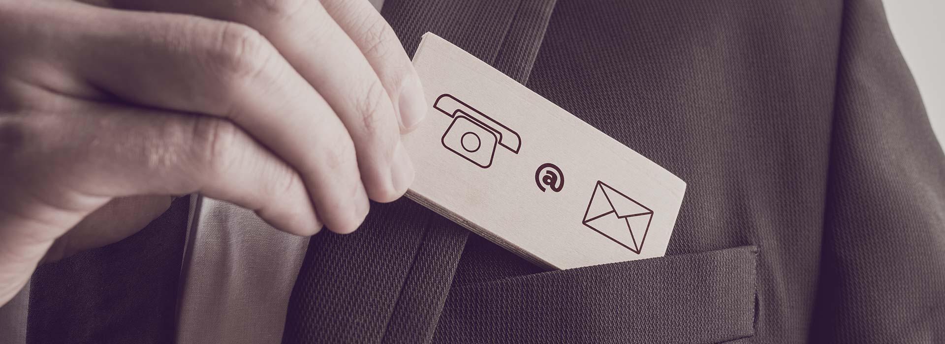 Eine Person im Anzug steckt eine hölzerne Visitenkarte in die Brusttasche eines Anzugs. Die Visitenkarte zeigt Symbole für Telefon, E-Mail und Post.