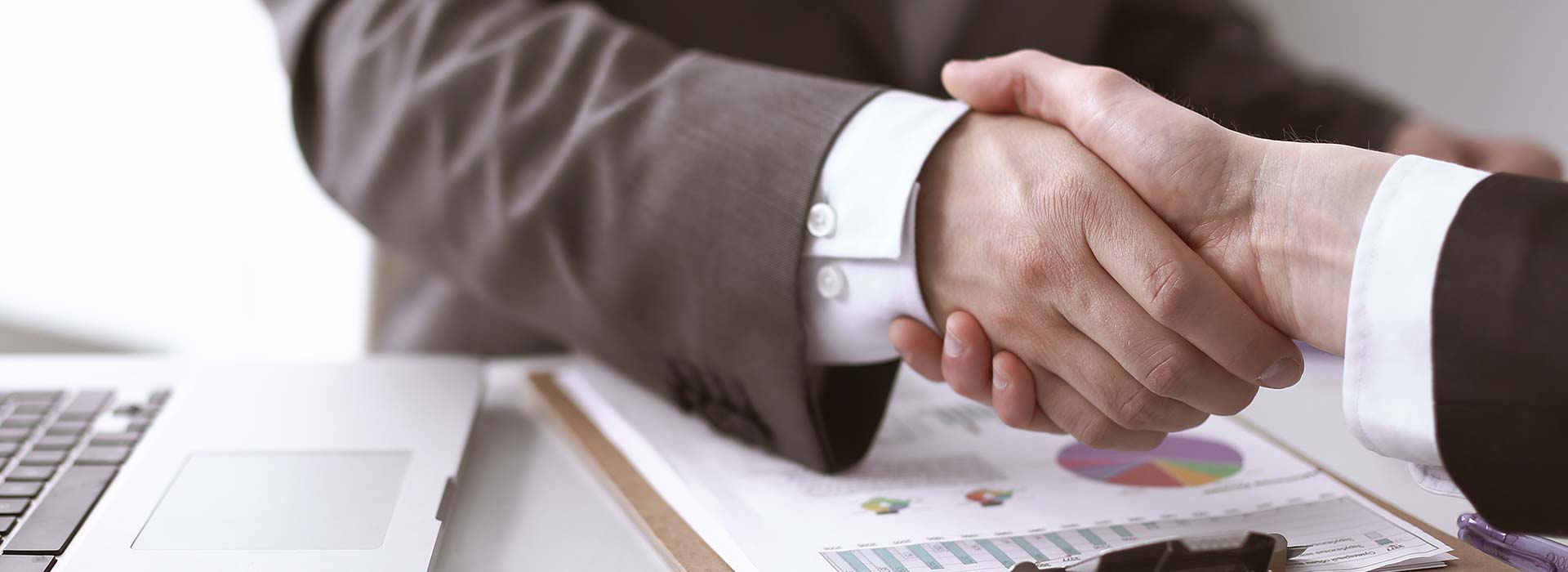Zwei Personen in Business-Kleidung geben sich die Hand. Links im Bild ist die Tastatur eines geöffneten Laptops zu sehen. Auf dem Schreibtisch liegt ein Klemmbrett mit Dokumenten, das oberste Dokument zeigt Diagramme.