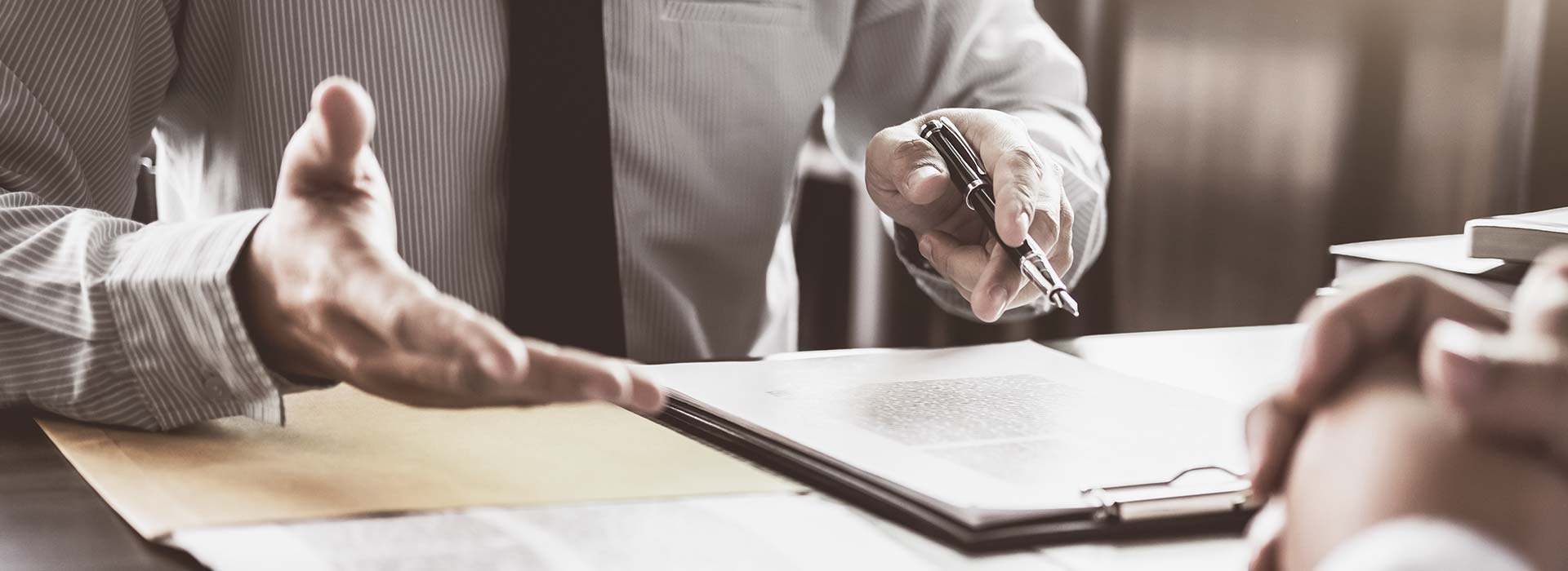 Ein Geschäftsmann in einem gestreiften Hemd und Krawatte sitzt an einem Tisch und führt ein Gespräch. Er gestikuliert mit einer Hand und hält in der linken Hand einen Stift über ein Dokument auf einem Klemmbrett. Im Vordergrund ist eine weitere Person mit verschränkten Händen zu sehen.